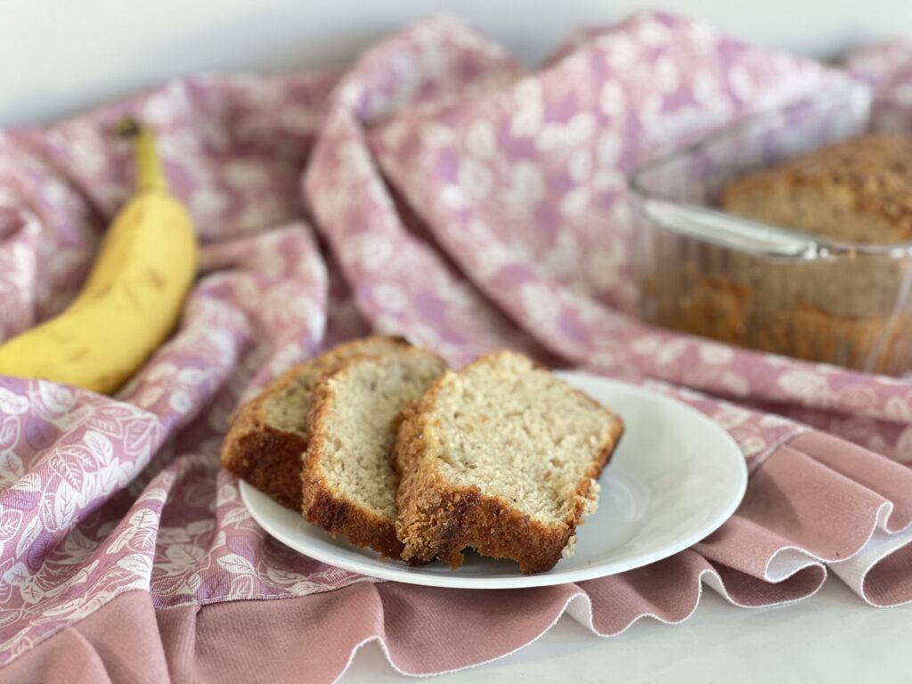three slices of banana bread on a small white plate in the foreground, with a yellow banana and glass pan with the remainder of banana bread loaf in the background. 